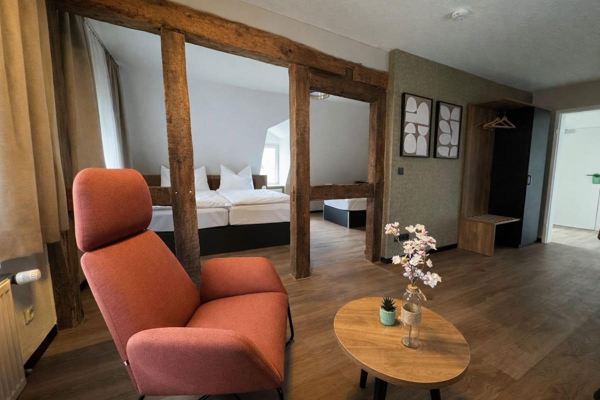 Una habitación moderna con un ventanal grande, una cama con estructura de madera, una silla roja, una pequeña mesa con un jarrón y un banco de madera.