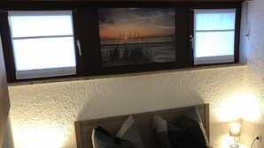 A bedroom with a bed, two bedside lamps, a nightstand, a window with a view, and a wall-mounted picture. - Vacation apartment Puustpaus close to the beach (Butjadingen)