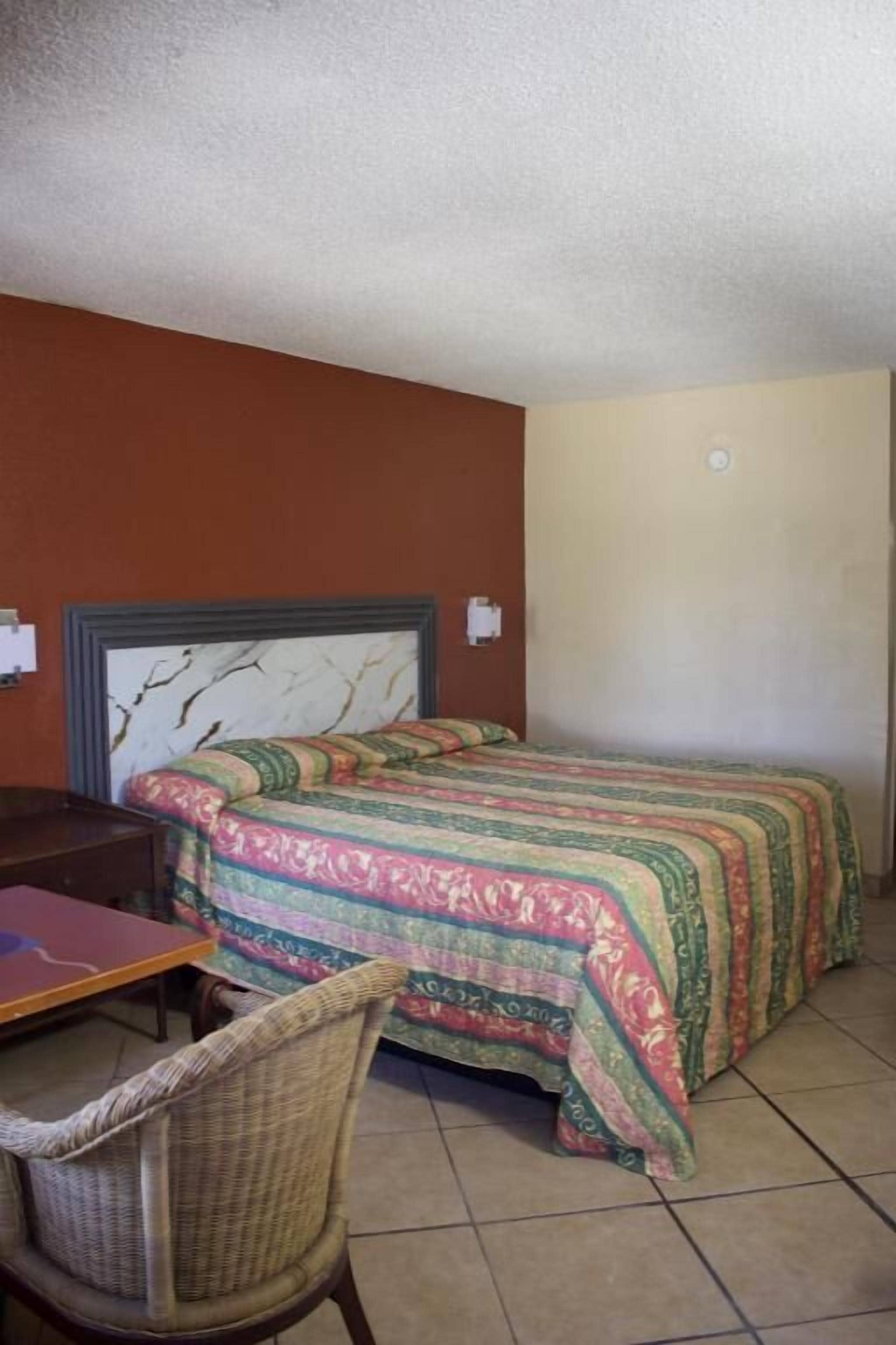 A single bed with a patterned bedspread, a wooden nightstand, a wicker chair, and a desk with a chair.