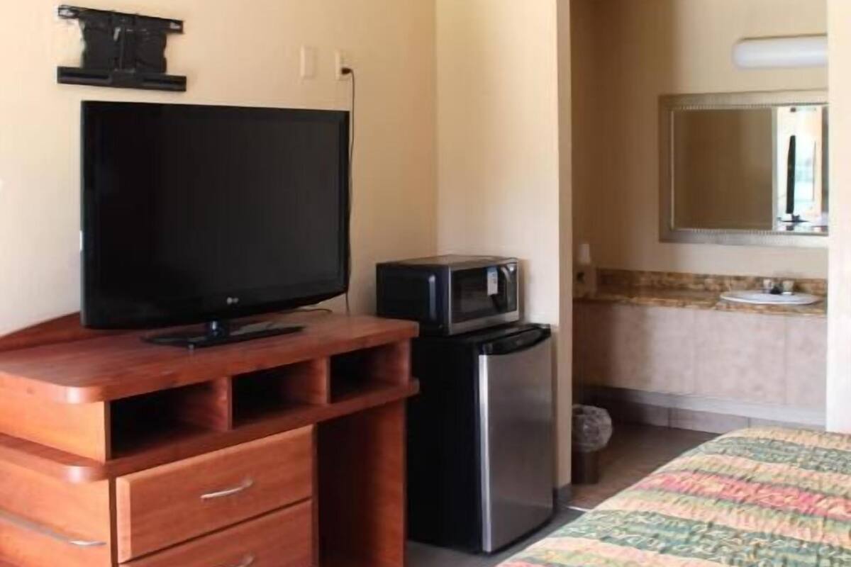 A hotel room with a wooden dresser, a flat-screen TV, a refrigerator, a bed with a patterned bedspread, and a bathroom with a sink and mirror.