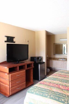 A hotel room with a wooden dresser, a flat-screen TV, a refrigerator, a bed with a patterned bedspread, and a bathroom with a sink and mirror. - Oak Ridge Inn (Kissimmee)