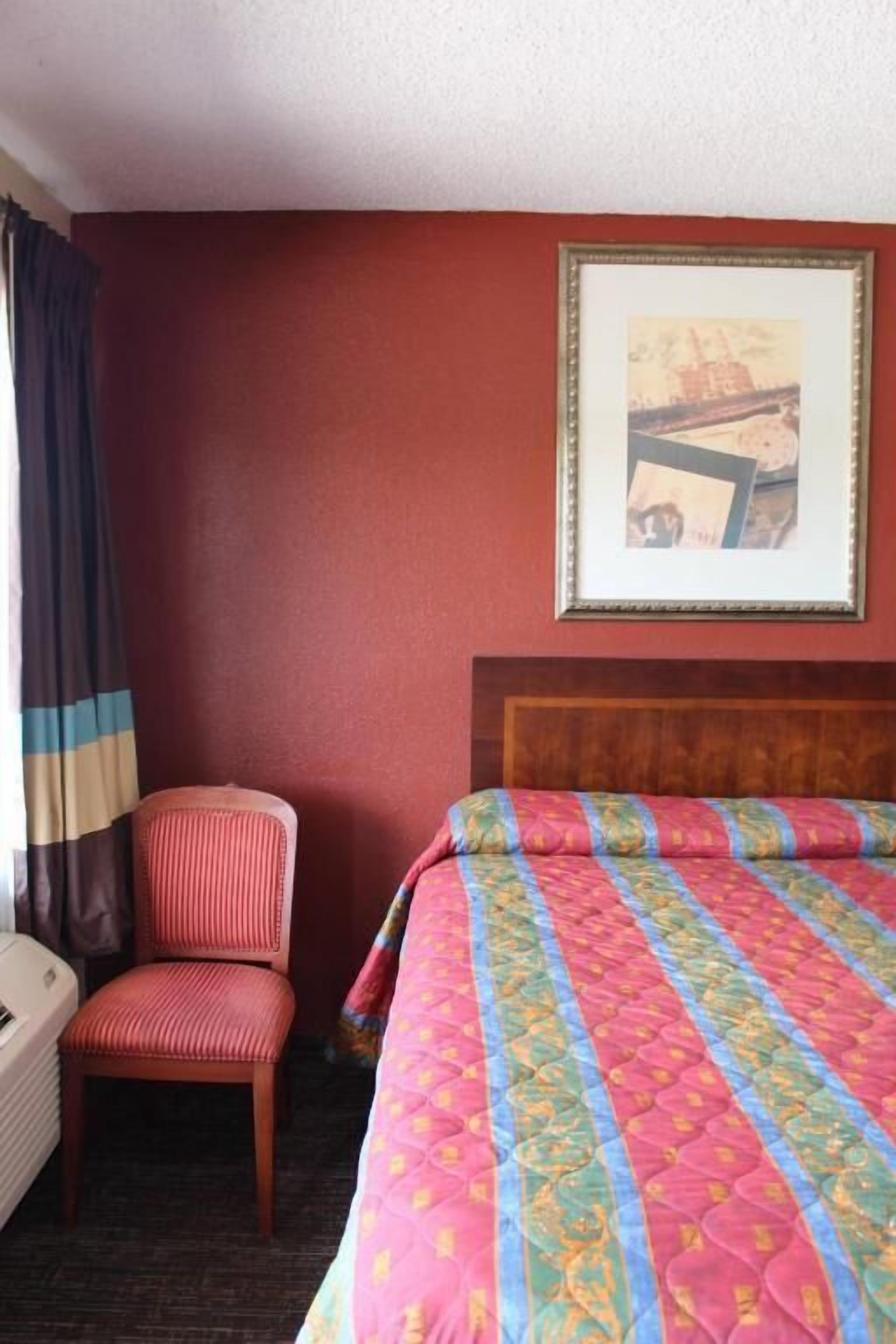Une chambre d’hôtel avec un lit, une chaise, un tableau encadré au mur et une fenêtre avec des rideaux.