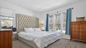 A bedroom with a tufted headboard, a bed with white bedding, a wooden dresser, a nightstand with a lamp, and a window with curtains. - St Paul Retreat:  Fireplace & Pet-Friendly (Saint Paul)