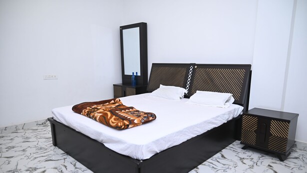 Een bed met een witte matras en een geborduurde deken, een zwart hoofdeinde, een nachtkastje met een spiegel en een vloer met een marmerprint.