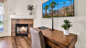 A room with a fireplace, a desk, a chair, and a large TV mounted on the wall. - Heritage Hideaway | 2BR Legacy Villa (La Quinta)