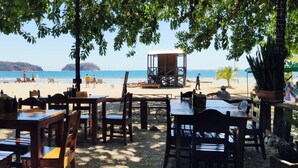 Un área de estar al aire libre con mesas y sillas de madera, una torre de salvavidas y vistas a la playa y al océano.