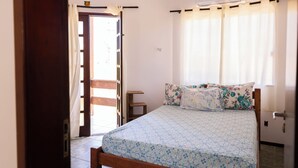 A bedroom with a bed, floral bedding, a small bedside table, and a window with curtains. - Casa com Hidro Piscina e Churrasqueira - Jacu pe (Camaçari)