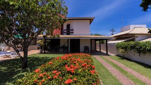 A two-story house with a covered patio, a swimming pool, and a well-maintained garden with red flowers. - Casa com Hidro Piscina e Churrasqueira - Jacu pe (Camaçari)