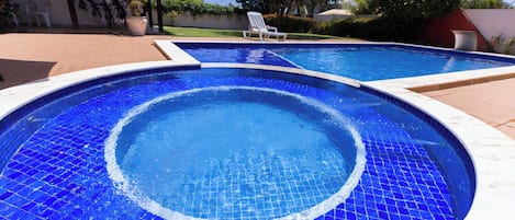 A swimming pool with blue mosaic tiles, surrounded by a patio area with chairs and a covered seating area.