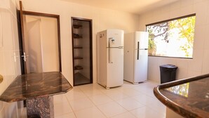 A kitchen with a marble countertop, two white refrigerators, a trash bin, and a window with a view of trees. - Casa com Hidro Piscina e Churrasqueira - Jacu pe (Camaçari)