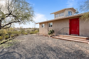 A two-story house with a red door, a small garden with rocks and a tree, and a clear sky.