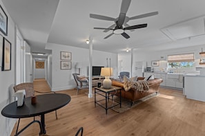 A spacious living room with a ceiling fan, a dining area with a table and chairs, a kitchen with a white countertop, and a large window.