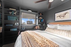 A bedroom with a large bed, a bookshelf, a window with a view, and a ceiling fan.