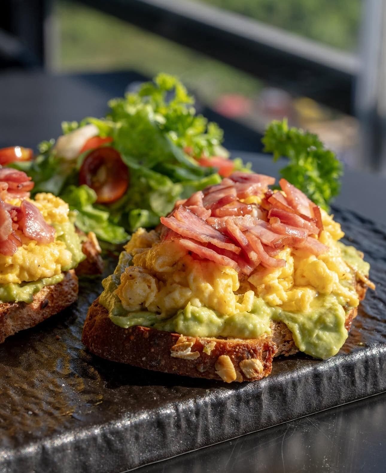 A breakfast dish with scrambled eggs, avocado, and bacon on toast, accompanied by a side salad.