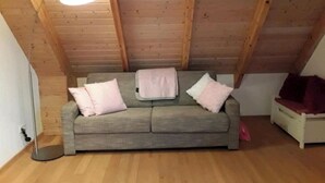 Quarto em estilo loft com teto de madeira, sofá cinza e mesa de cabeceira pequena e branca.