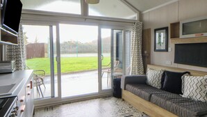 A modern kitchen with a stove, a dining table, and chairs. There is a sliding glass door leading to a patio with a lawn and chairs. - The Family Pod - Gower (Swansea)