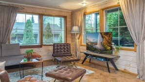 Interior acolhedor de uma cabana com sofá, um banquinho de couro, uma televisão sobre um suporte e uma janela com cortinas.