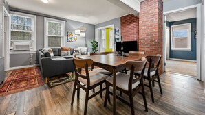 A modern living room with a grey sofa, wooden dining table, and chairs. There is a brick fireplace, a large window, and a door leading to another room. - Spacious 5BR Retreat - Duplex close to Attitash (Bartlett)