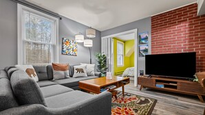 A modern living room with a sectional sofa, a flat-screen TV, and a brick accent wall. - Spacious 5BR Retreat - Duplex close to Attitash (Bartlett)
