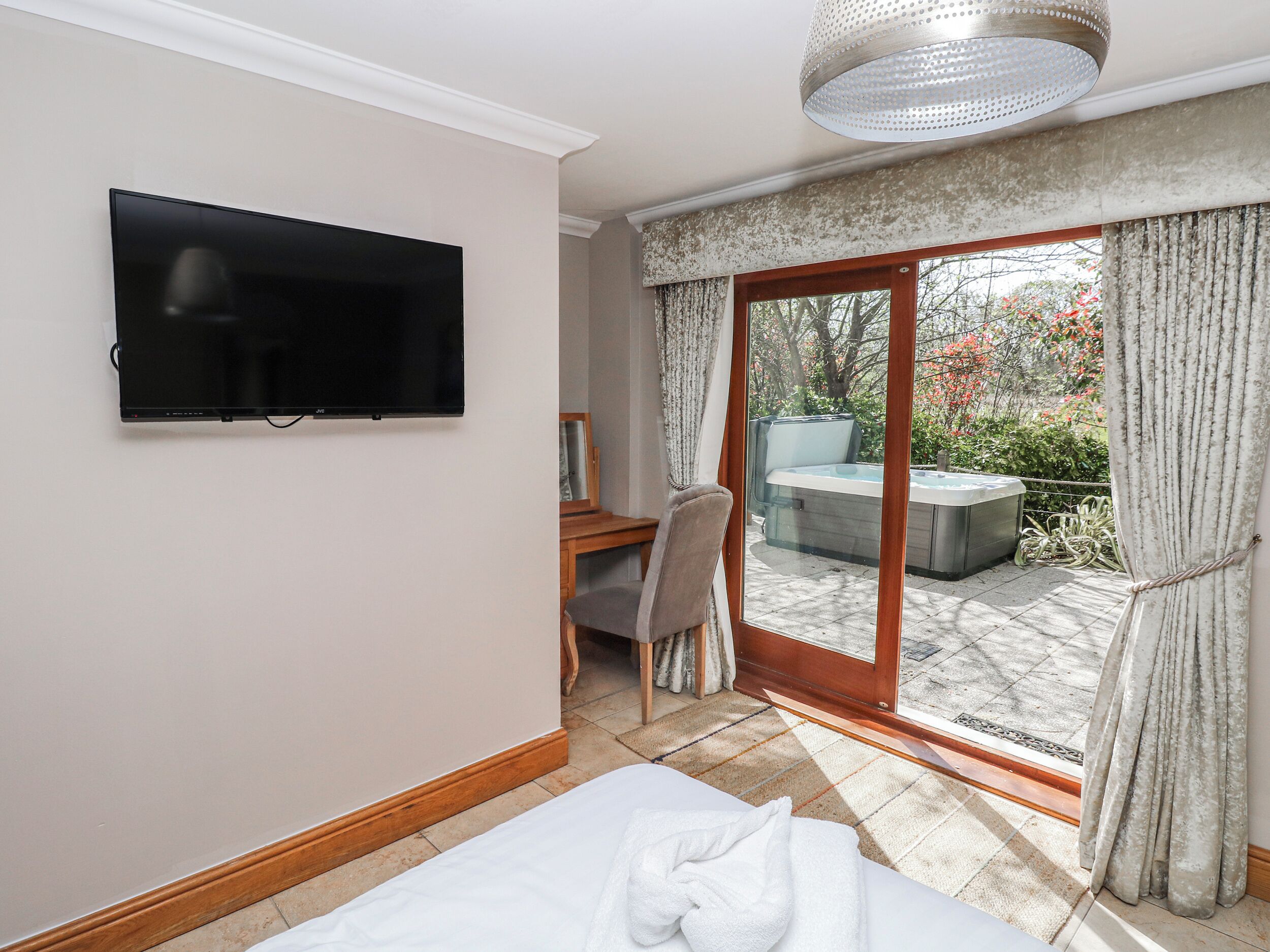 A hotel room with a flat-screen TV mounted on the wall, a bed with a white comforter, a desk with a chair, and a sliding glass door leading to an outdoor hot tub area.