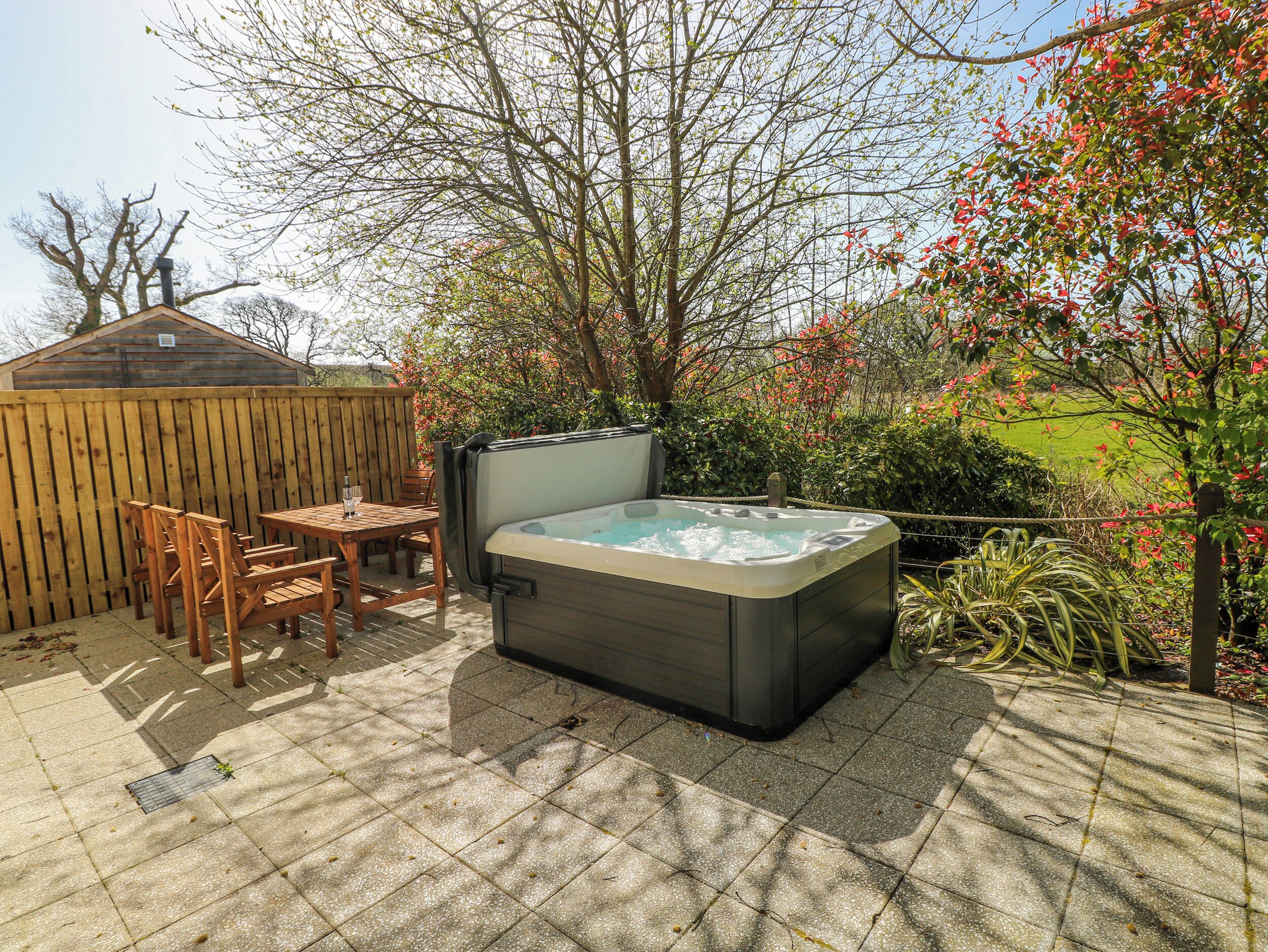 Un jacuzzi en un patio trasero con un cerco de madera, una mesa con sillas y una construcción de madera al fondo.