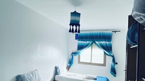 Ein Zimmer mit zwei Betten, einem Fenster mit blauen Vorhängen, einer Pendelleuchte und einem Ventilator.