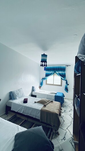 A room with two beds, a window with blue curtains, a hanging light fixture, and a fan. - Spacious apartment with terrace - 2 bedrooms, 5 beds, fully equipped (Chefchaouen)