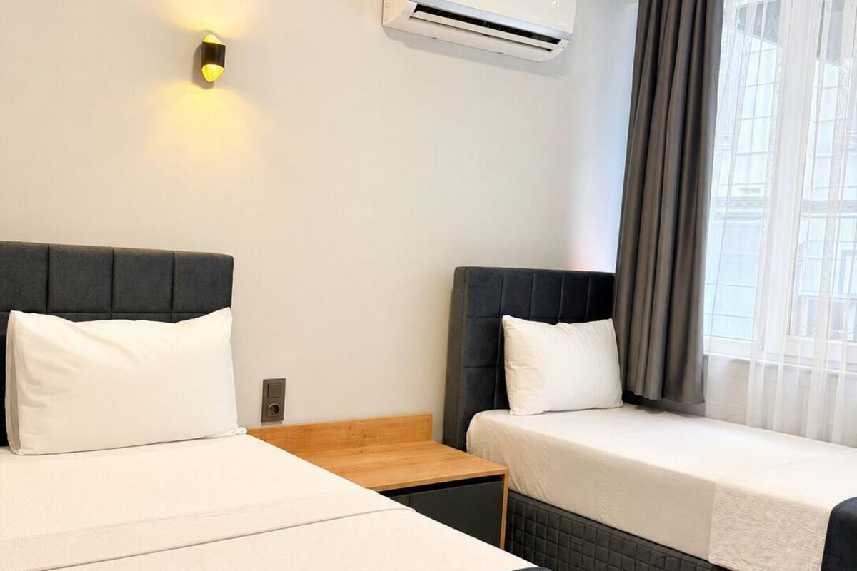 Une chambre d’hôtel avec deux lits, une table de chevet, un appareil de climatisation, une lampe fixée au mur et une fenêtre avec des rideaux.