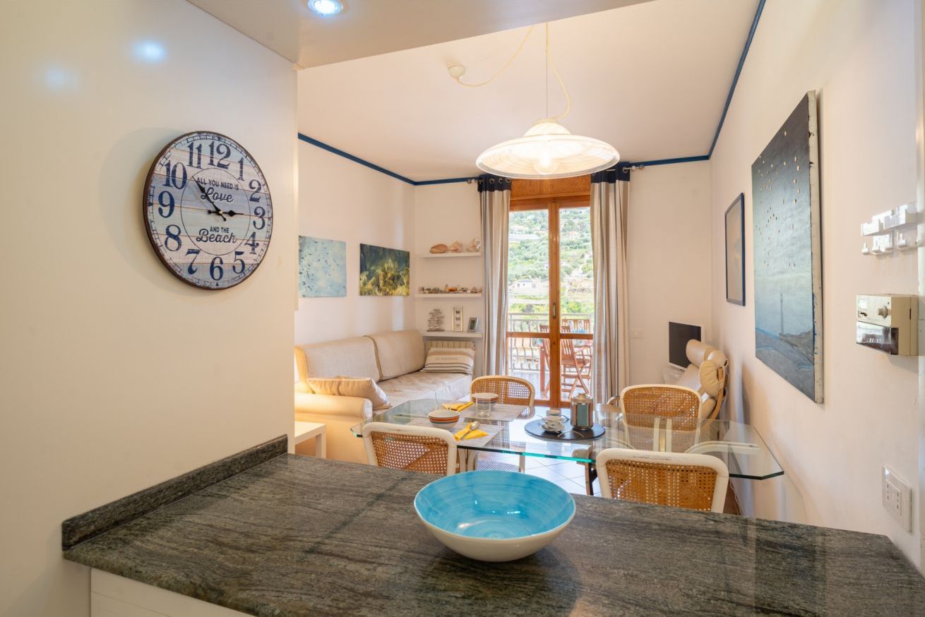 A modern kitchen with a large clock, a dining area with wicker chairs, and a view through a glass door.