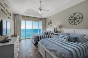 Ein Schlafzimmer mit einem großen Bett, zwei Einzelbetten, einem Deckenventilator, einem Fernseher und Blick auf den Ozean.