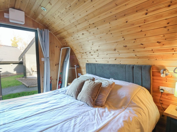 A bedroom with a wooden ceiling, a bed with white bedding, a grey headboard, and a window with curtains. - Glamping Pod 4 - Gower (Swansea)