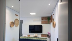 A modern bedroom with a flat-screen TV mounted on the wall, a bed with a plaid bedspread, and a small bedside table with a lamp. - Skyline Confort Club - Araucaria - Penha/sc (Penha)