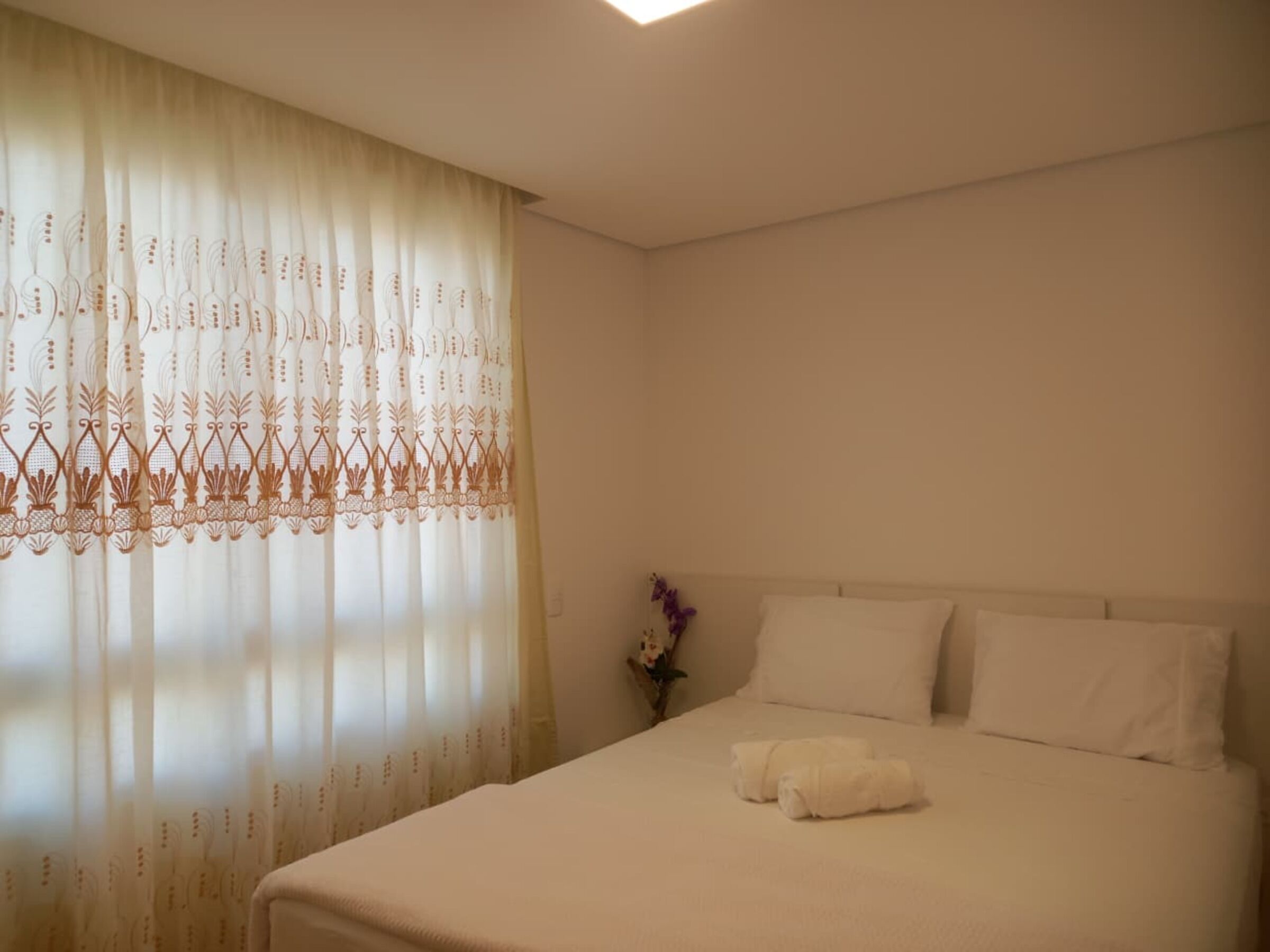 A neatly made bed with white linens and two rolled towels, a single purple orchid, and a window with sheer curtains featuring a decorative pattern.
