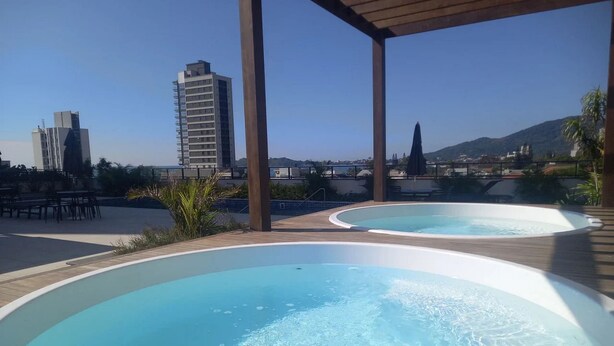 A rooftop pool area with two hot tubs, wooden deck, and a view of city buildings and hills. - Skyline Confort Club - Araucaria - Penha/sc (Penha)