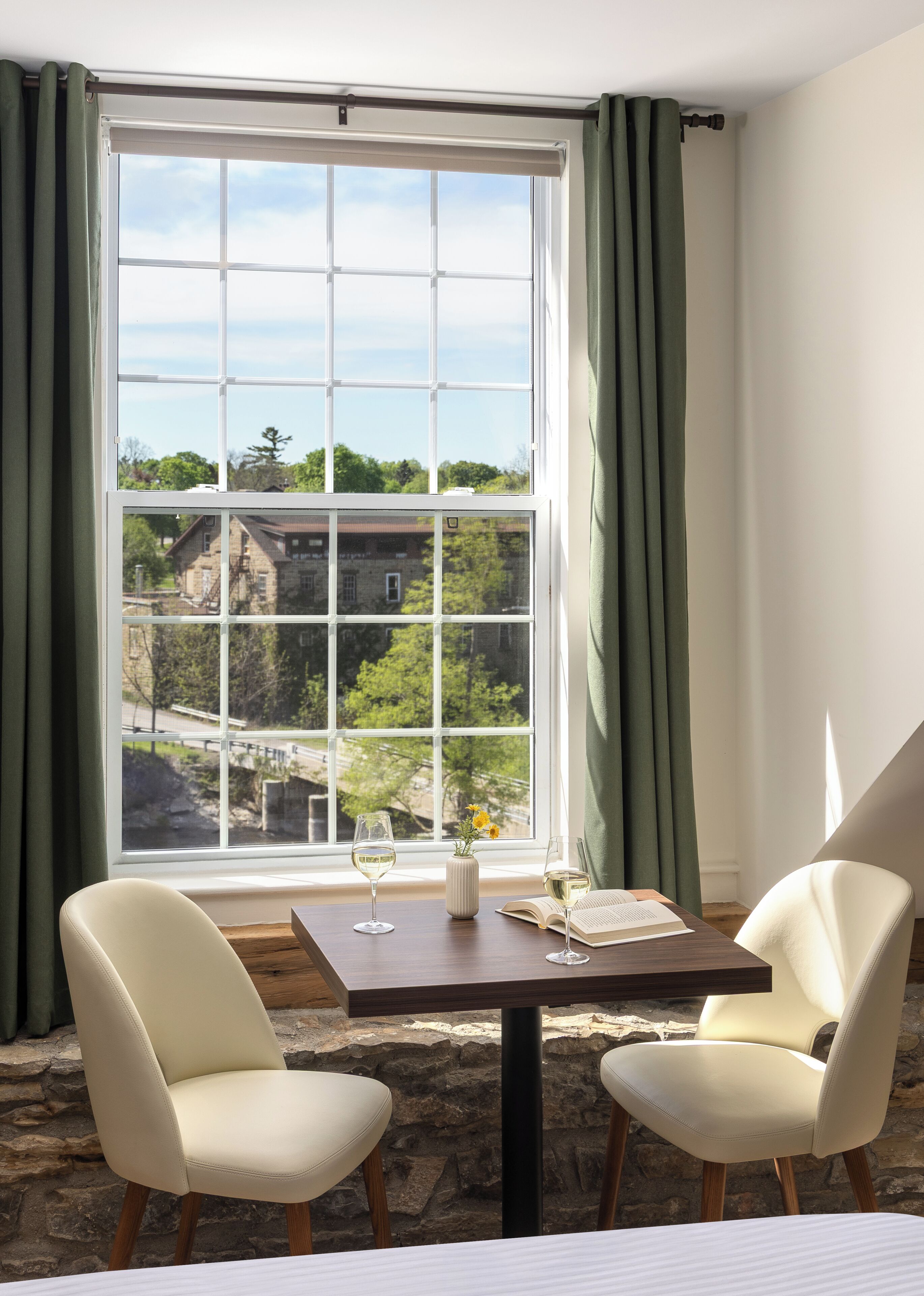 Una habitación con una mesa, dos sillas, un ventanal con cortinas y vistas a un edificio y árboles.