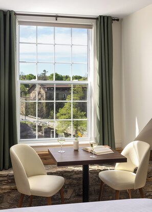 A room with a table, two chairs, a window with curtains, and a view of a building and trees. - Tri Century Mill Suites (Carleton Place)