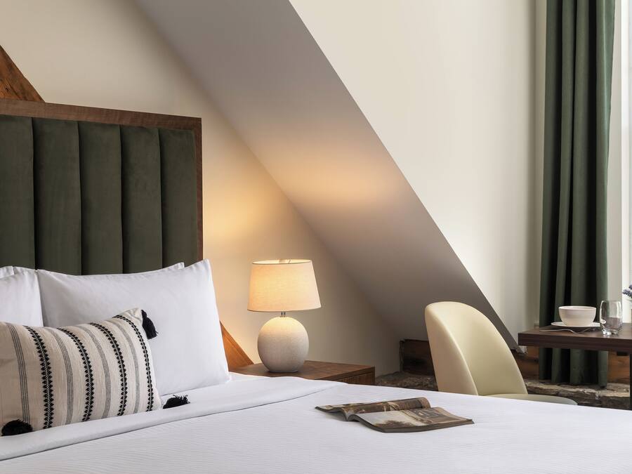 Habitación de hotel con cama, mesita de noche, lámpara, silla y ventana con cortinas.