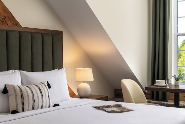 A hotel room with a bed, a nightstand, a lamp, a chair, and a window with curtains. - Tri Century Mill Suites (Carleton Place)