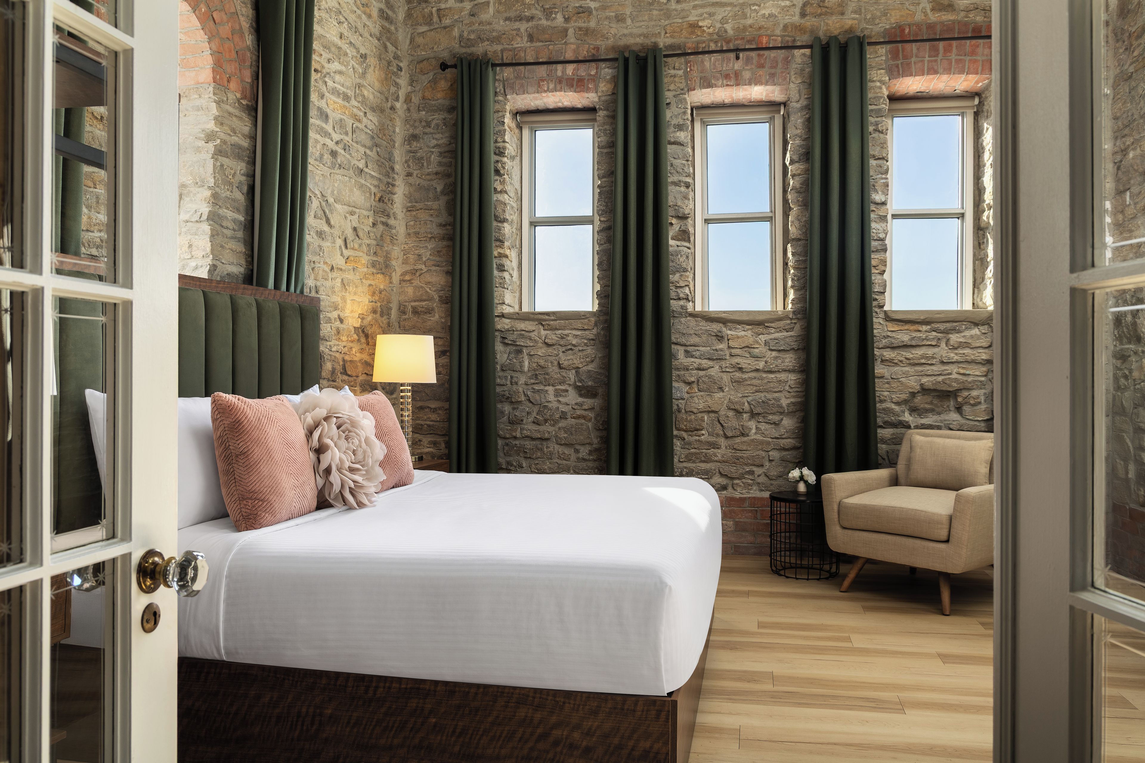 A bedroom with a stone wall, a bed with white bedding, a green headboard, and a small table with a lamp.