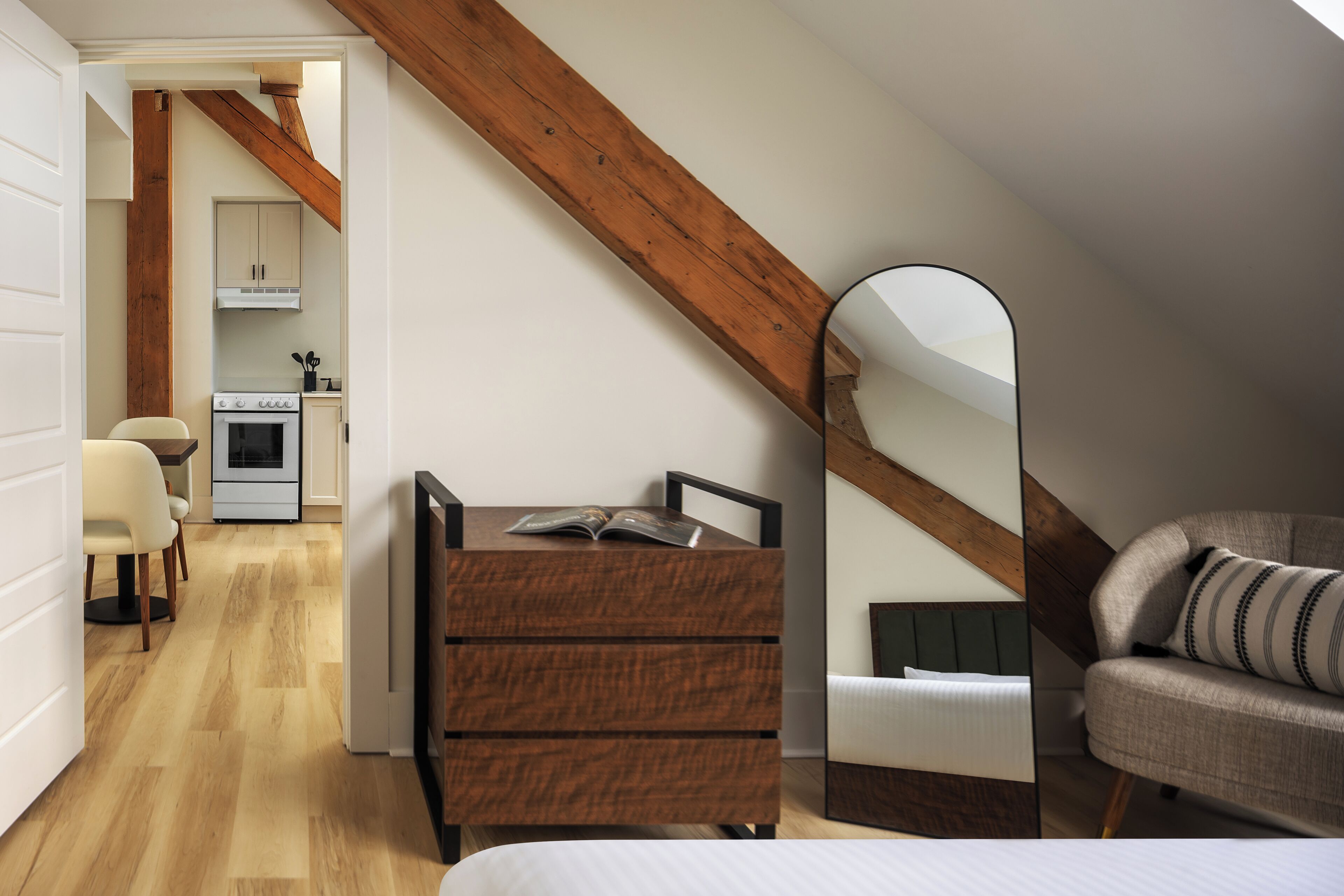 A bedroom with a wooden ceiling, a bed, a dresser, a mirror, and a view of a kitchen and dining area.