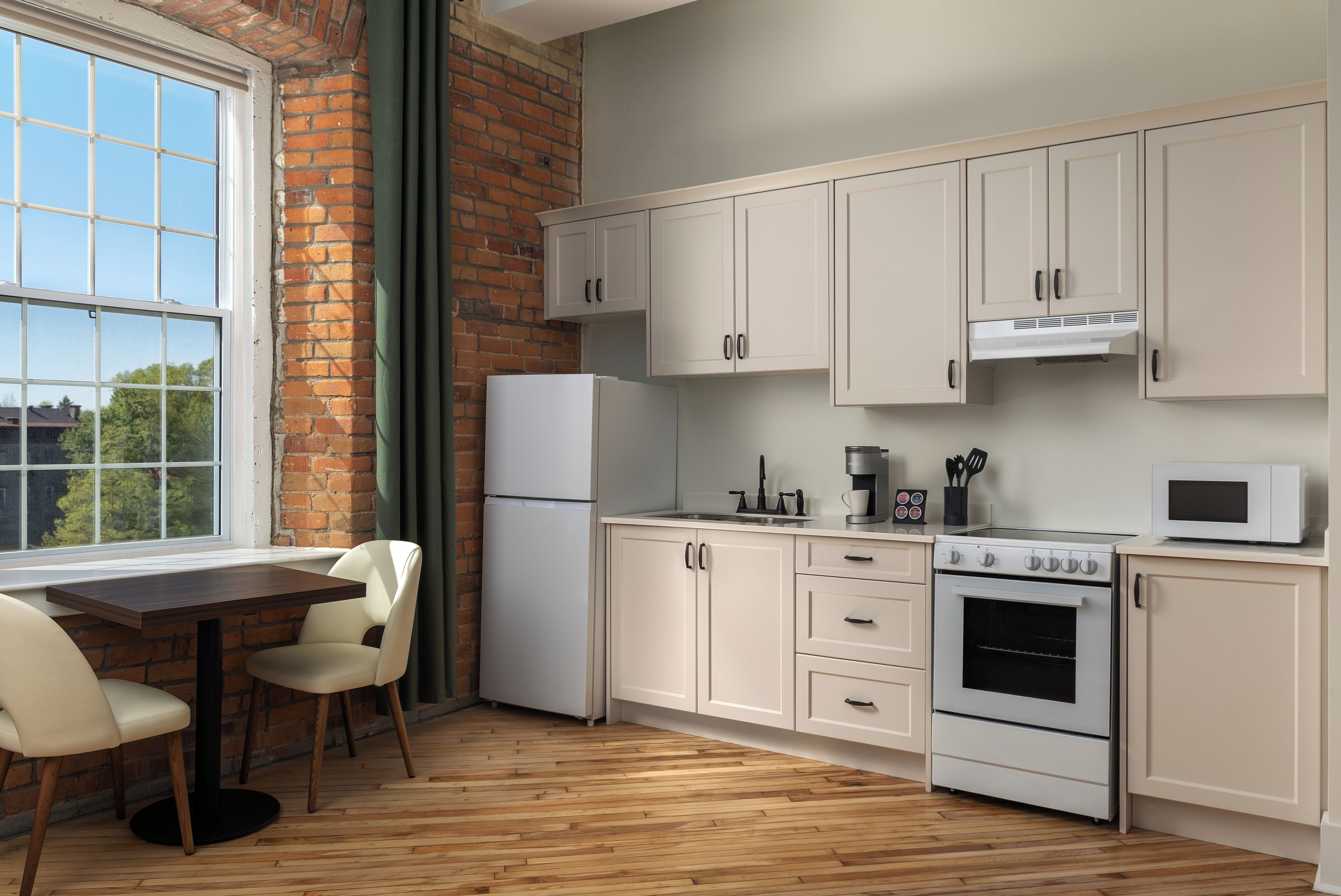 Una cocina con armarios blancos, un refrigerador, una estufa y un microondas. Hay una ventana con vistas a árboles y un muro de ladrillo.
