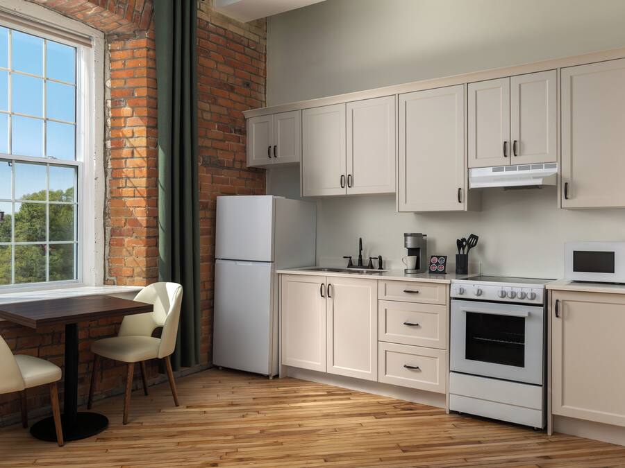 Una cocina con armarios blancos, un refrigerador, una estufa y un microondas. Hay una ventana con vistas a árboles y un muro de ladrillo.