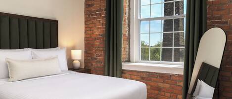 A hotel room with a bed, a nightstand, a lamp, a window with a view of a brick building, and a mirror.