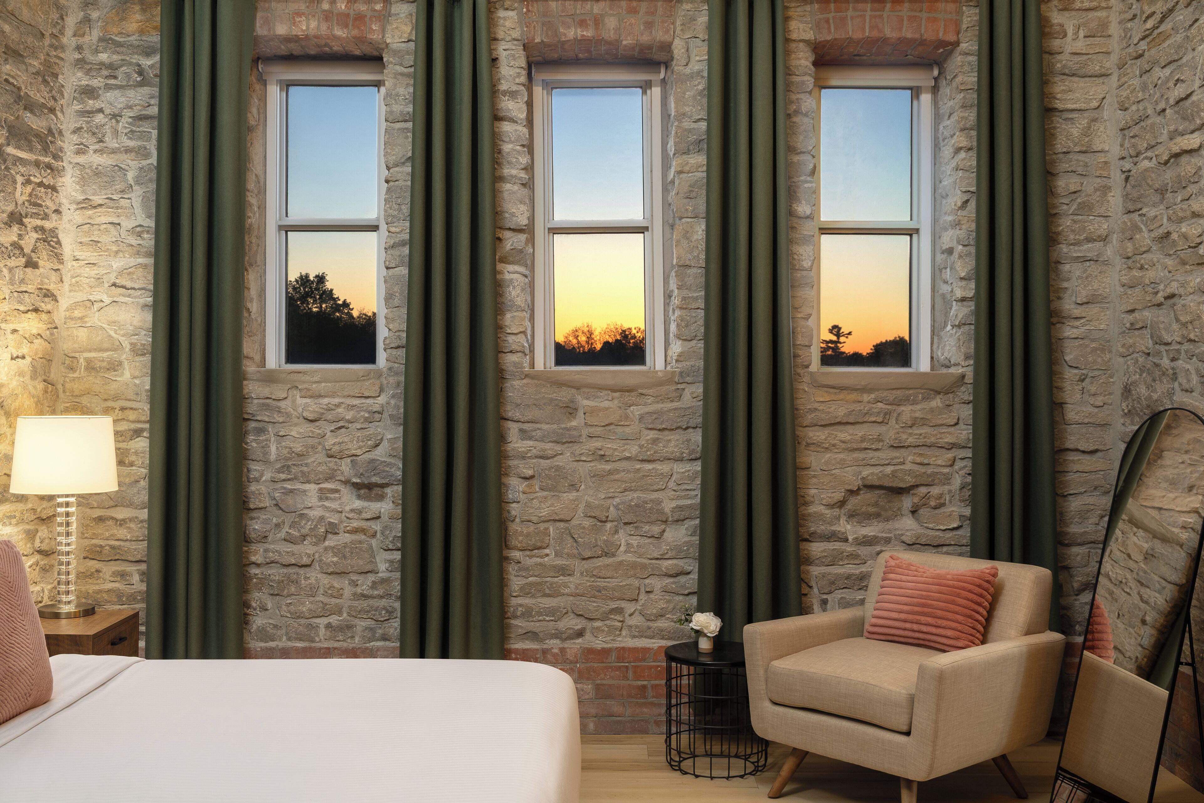 Una habitación con pared de piedra, tres ventanas con cortinas, una cama, una silla, una lámpara y una mesita con un florero.