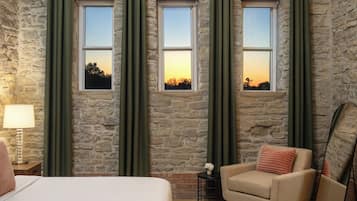 Una habitación con pared de piedra, tres ventanas con cortinas, una cama, una silla, una lámpara y una mesita con un florero.