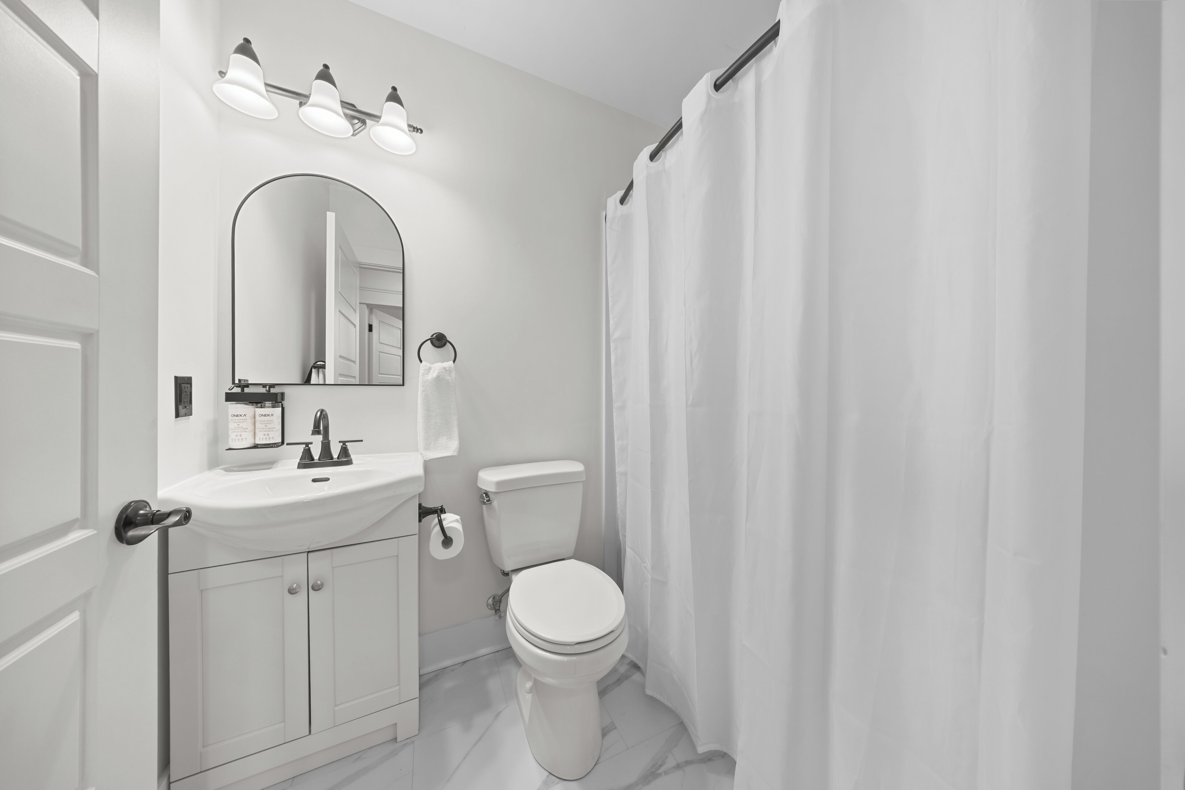 Un baño con lavamanos blanco, espejo y retrete. Hay tres luces sobre el espejo y una cortina de ducha.