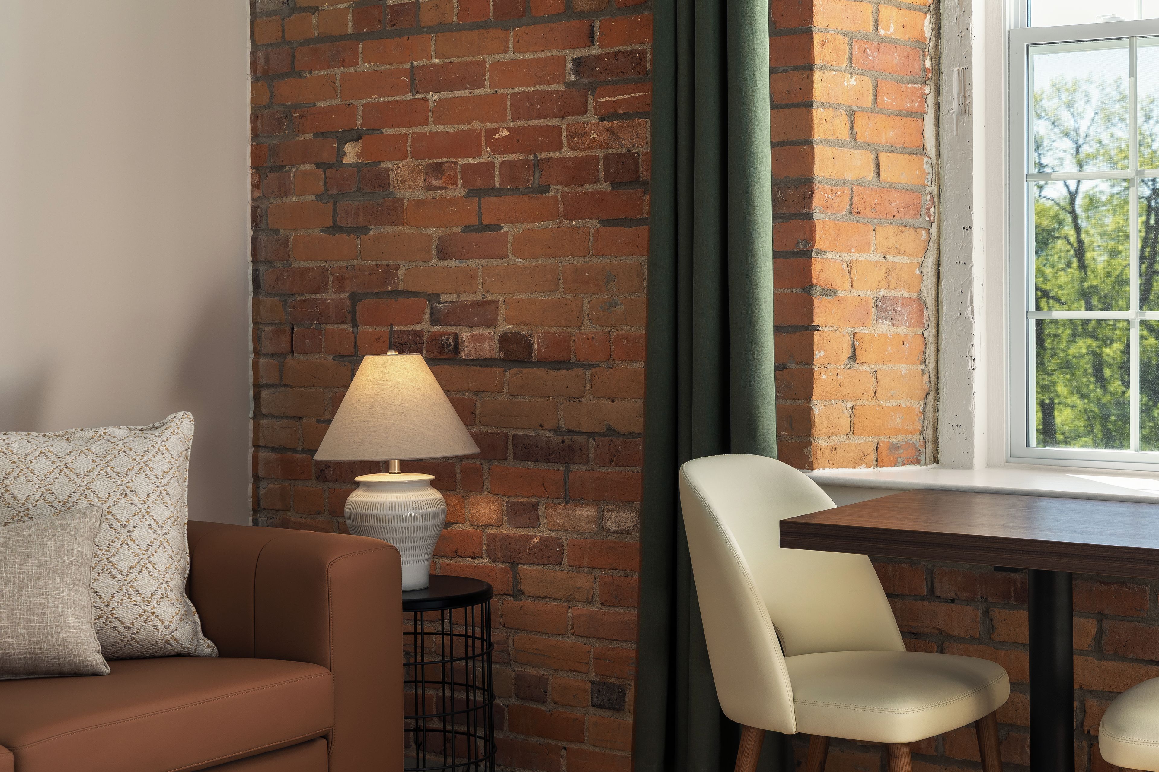A room with a brick wall, a table, a chair, a lamp, and a window with a view of trees.