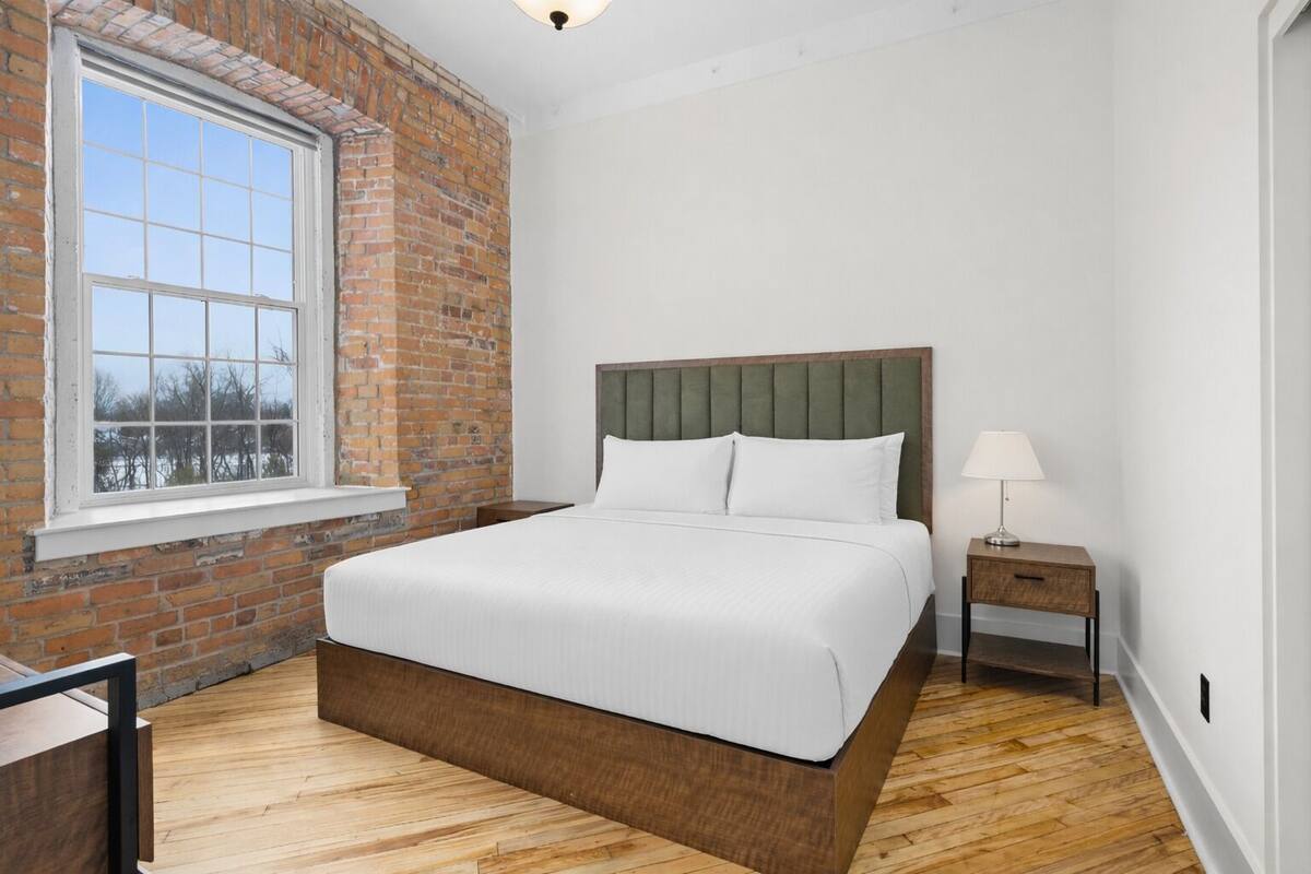 A bedroom with a brick wall, a large window, a wooden floor, a bed with white bedding, and a nightstand with a lamp.