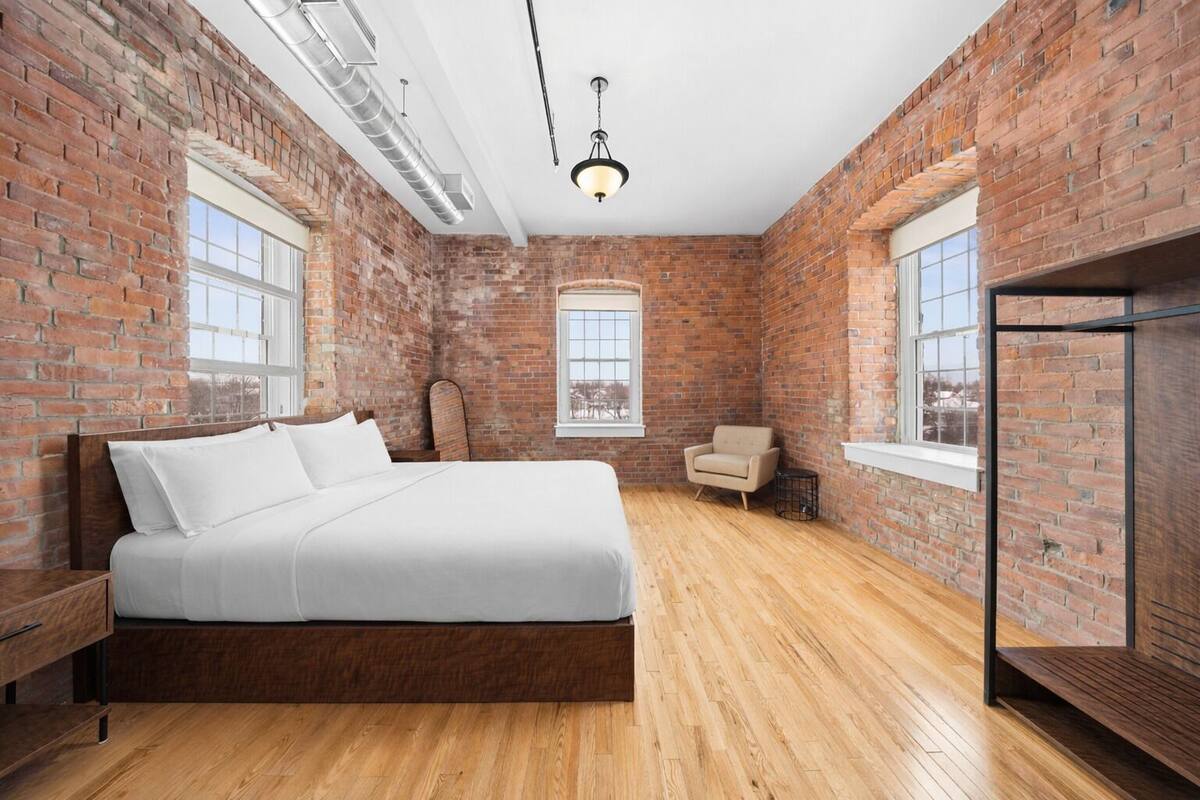 A bedroom with a brick wall, a wooden bed, a chair, and a window.