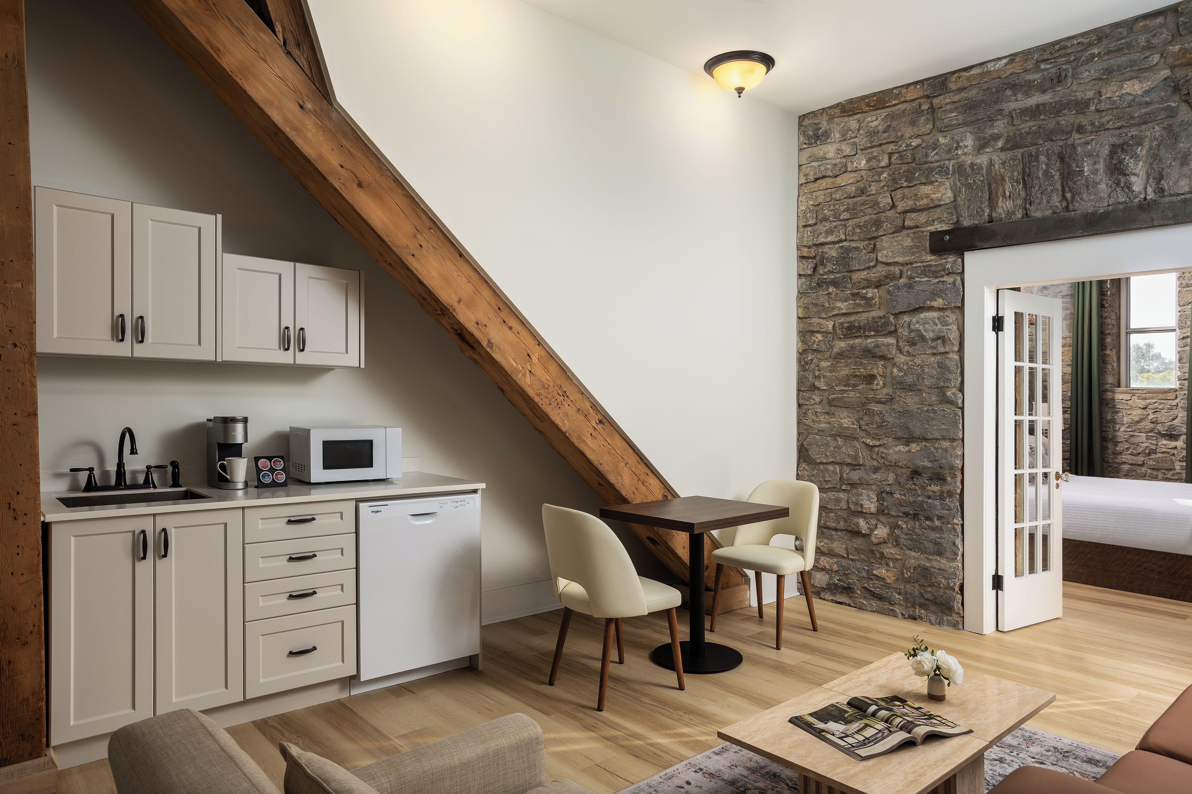 Cocina moderna con armarios blancos, microondas y cafetera. Hay una zona de comedor con mesa y sillas, y una pared de piedra al fondo.